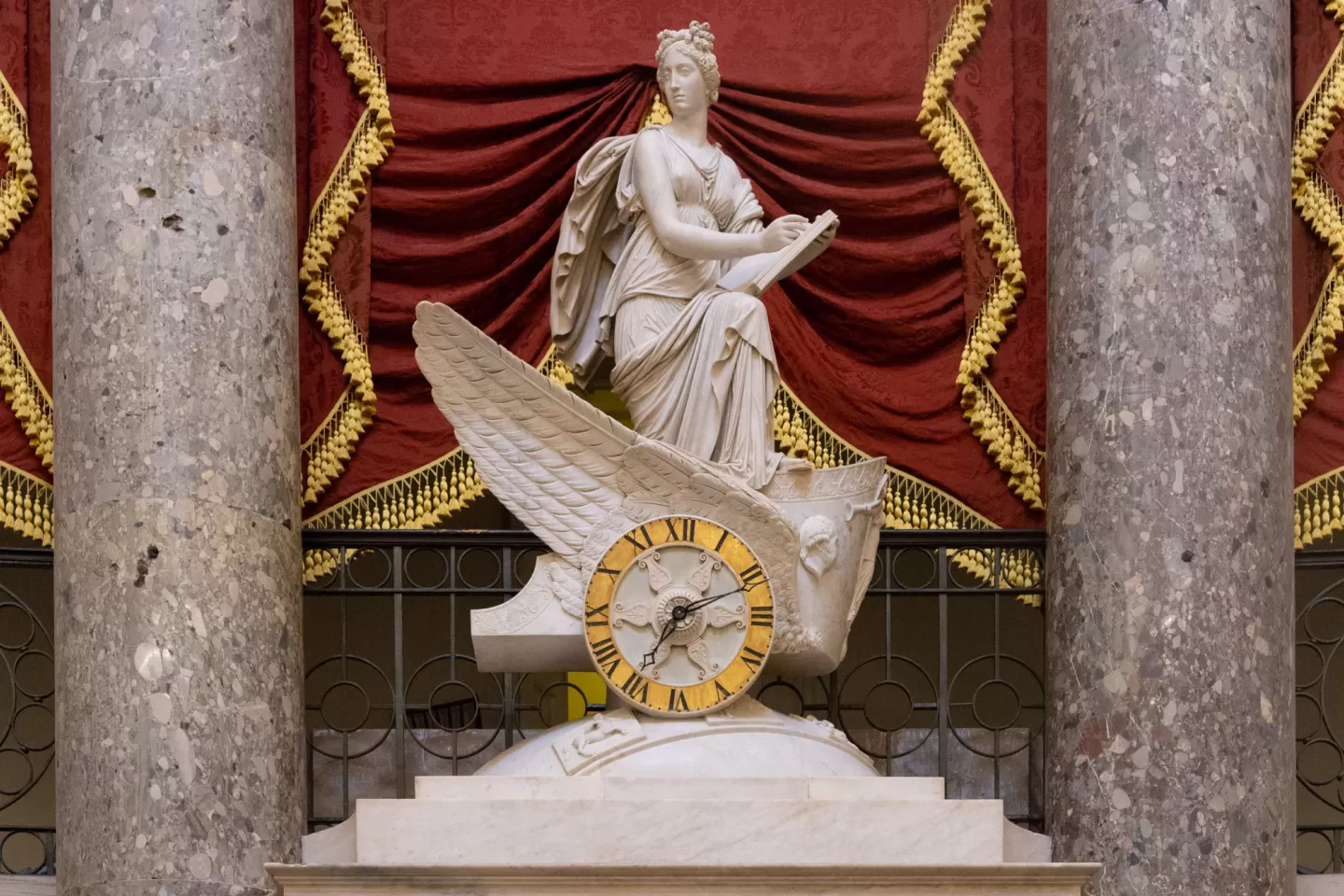 Marble statue between two columns with red drapery behind.