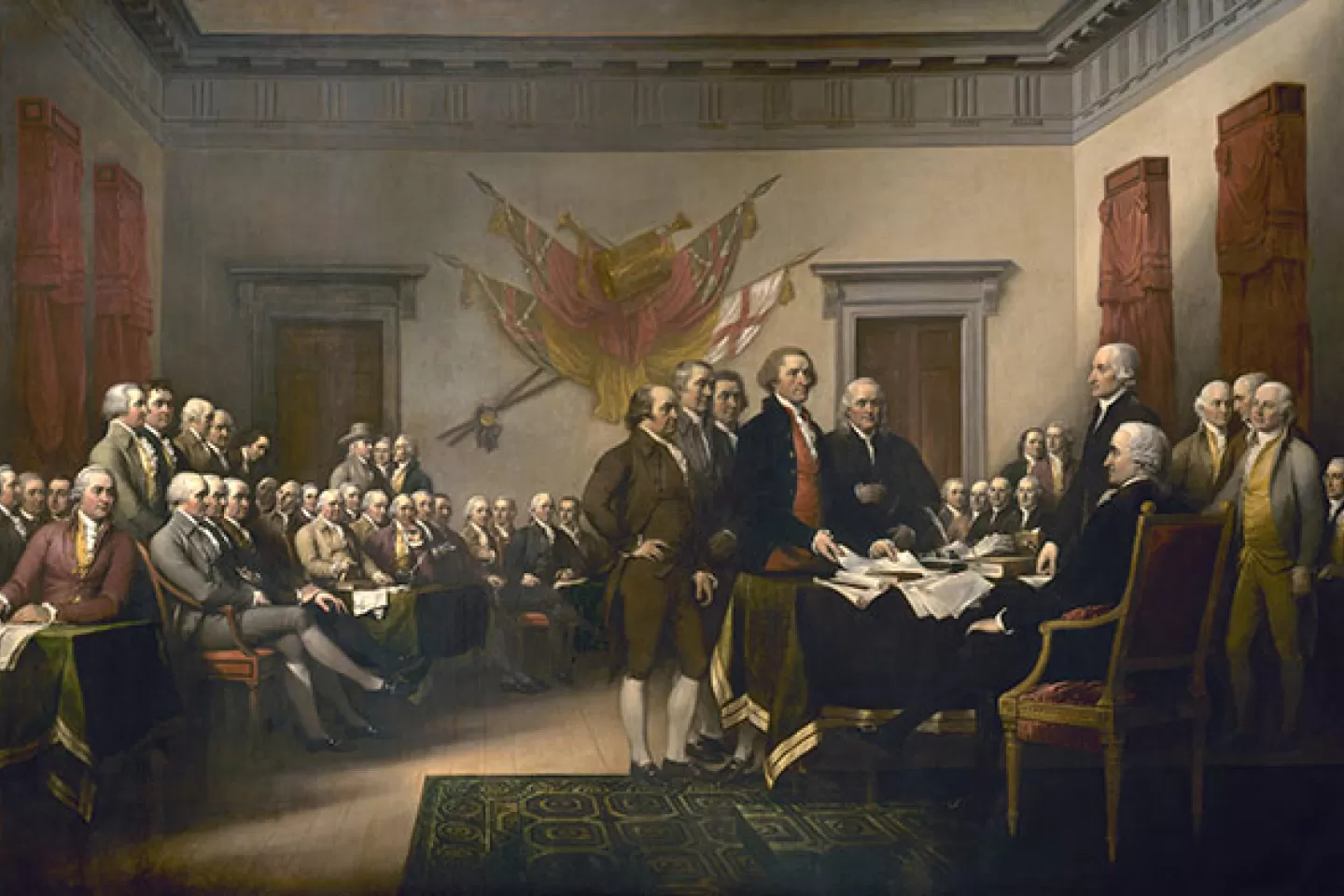 Trumbull's painting of the signing of the Declaration of Independence. Many men in suits stand in an office gathered around a desk. 