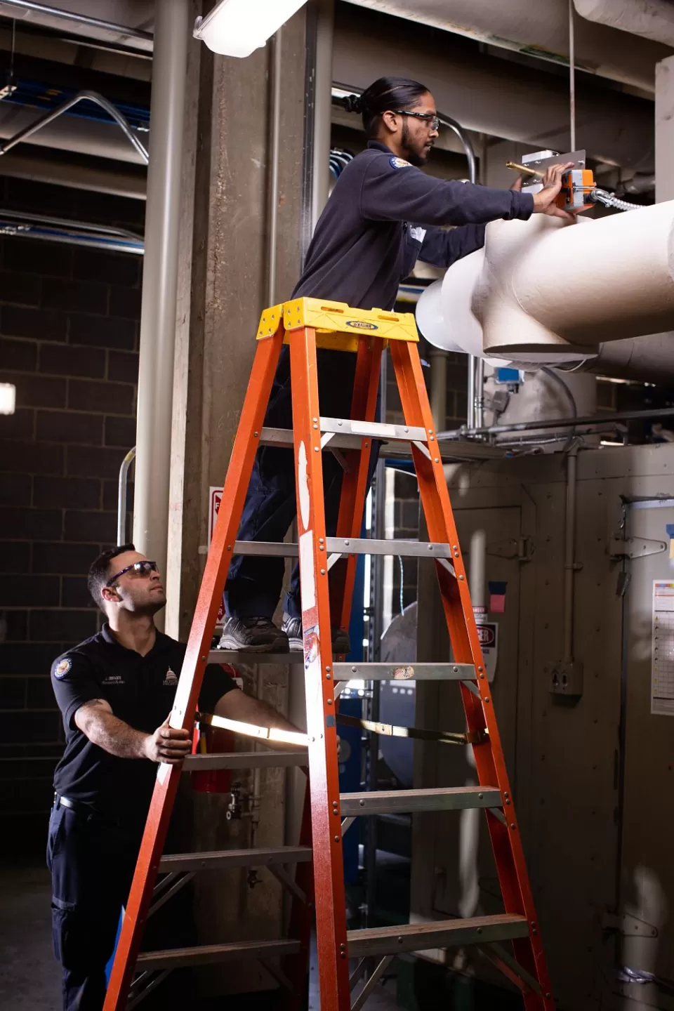 A/C Equipment Mechanic Anthony Mungal replaces  a malfunctioning chilled water valve actuator, while Justin Zibragos assists by steadying the ladder.