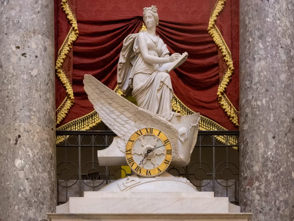 Marble statue between two columns with red drapery behind.