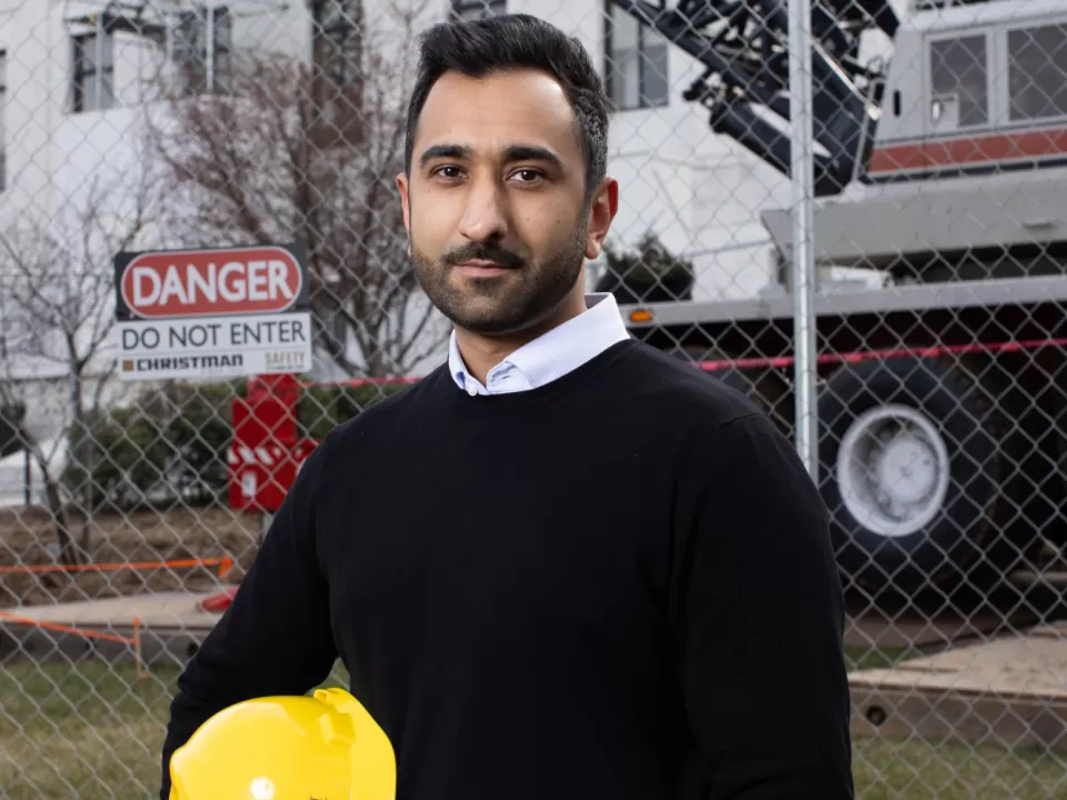 Person standing holding a yellow hardhat with a fence and construction equipment behind.