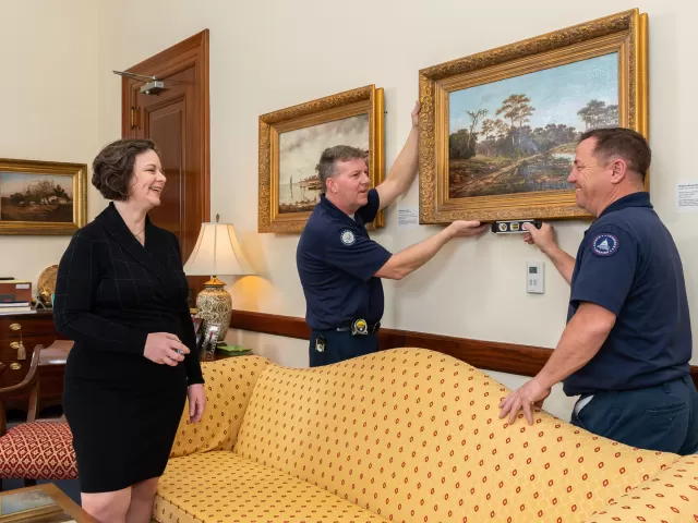 Senate Move Coordinator Bonnie Holod, who is also the supervisory architect in the Senate Office Buildings jurisdiction, works with Michael Gass and Paul Bosch, wood crafters with the Senate Wood Crafting Branch, to hang artwork in a senator's office.